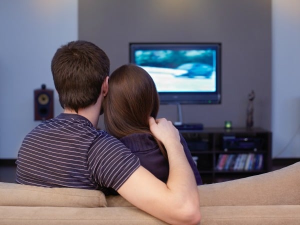 a husband with his arm around his wife while they sit on the couch watching tv