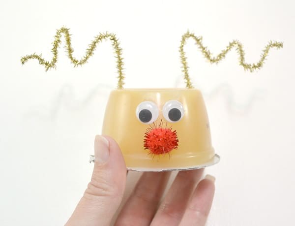 a hand holding an upside down applesauce cup with googly eyes, a red pom pom, as a nose, and gold pipe cleaners as antlers on it on a white background