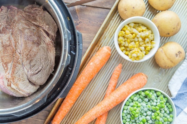 beef roast cooking in an instant pot next to carrots, potatoes, corn and peas on a wood tray