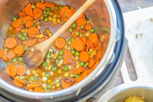 carrots, peas, corn, sauce and a wooden spoon in an instant pot next to a white cutting board