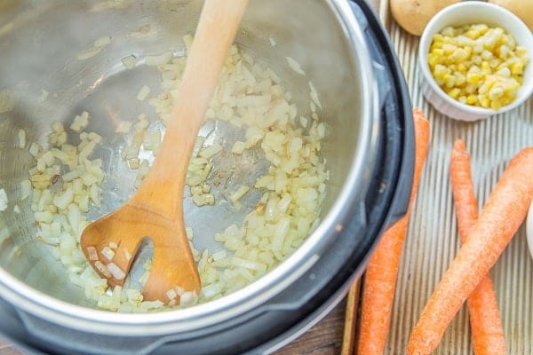 olive oil, diced onions, minced garlic cooking in an instant pot with a wooden spoon, next to a tray of carrots, frozen corn and potatoes