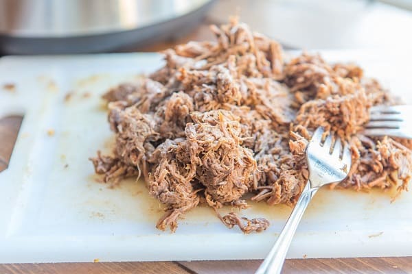 two forks and shredded beef on a white cutting board on a wood table with an instant pot in the background