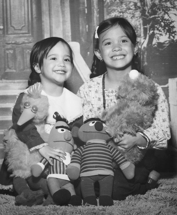 two young girls smiling as they have their picture taken while holding Sesame Street stuffed animals against the backdrop of a house