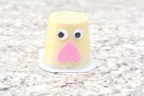 an upside down vanilla pudding cup with two googly eyes and an upside down pink foam heart on it on a gray counter