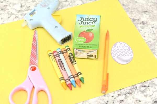 yellow construction paper, scissors, hot glue gun, a pencil, a juice box, crayons, and a printable Easter egg on a counter