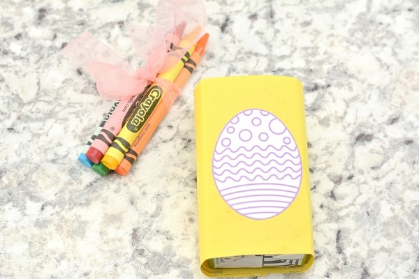 a juice box wrapped in yellow construction paper with a printed white easter egg with decorative lines on it next to some crayons tied together with an orange ribbon on a gray kitchen counter