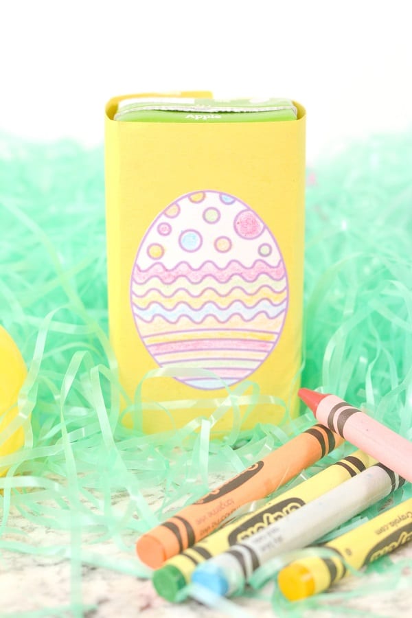 a juice box wrapped with yellow paper with a white paper egg colored in on it, on fake green grass next to a plastic egg and crayons