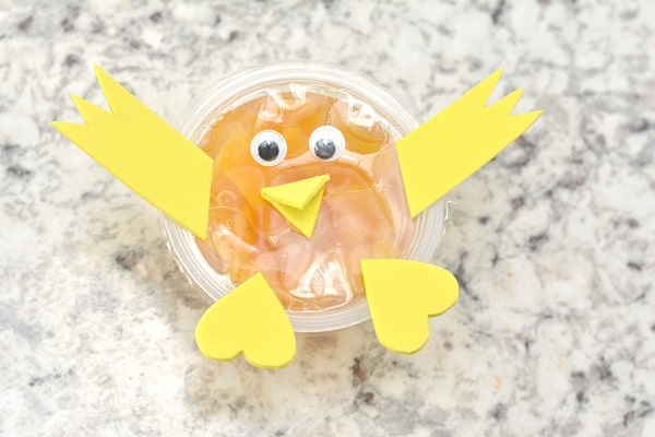 a peach fruit cup with googly eyes and a yellow foam beak, wings, and heart shaped feet on it on a gray counter