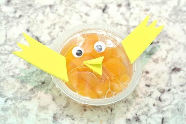 a peach fruit cup with googly eyes and a yellow foam beak and wings on it on a gray counter