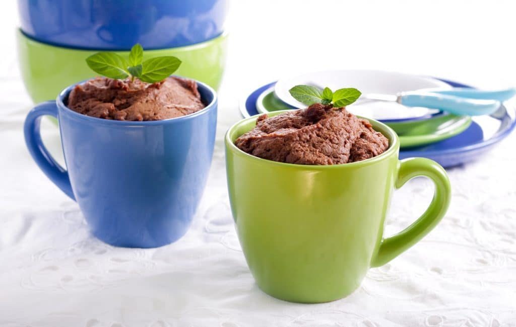 two microwave mug desserts in blue and green mugs with blue and green plates and bowls in the background, all on a white cloth