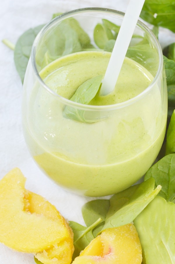 peachy green smoothie in a glass with a straw in it next to peach slices and spinach leaves on a white background