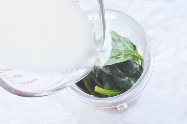 milk being poured from a glass measuring cup into frozen peach slices and spinach leaves in a cup on a white background