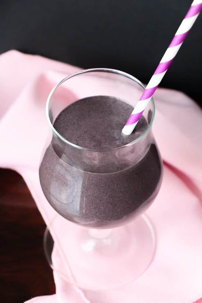 a blueberry spinach smoothie in a glass with a pink and white straw on a pink cloth on a black background