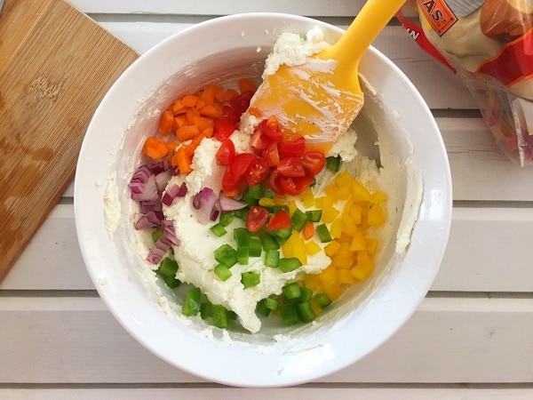 cream cheese, bell peppers, carrots, red onions, and cherry tomatoes in a white bowl with a yellow spatula on a white wood table with a wooden cutting board and a bag of bagels in the background