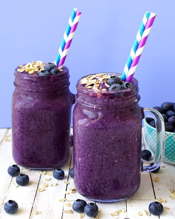 two blueberry muffin smoothies in glasses with blue and white, and purple and white striped straws, with more blueberries and oats on a wood table with a blue background