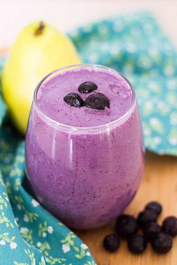 blueberry pear smoothie topped with three blueberries in a glass next to a blue flower cloth, a pear, and blueberries on a brown table 
