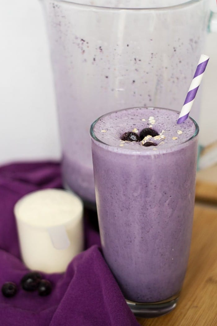 blueberry protein smoothie in a glass topped with blueberries with a purple and white straw in it next to a purple cloth on a brown table with a blender, blueberries and plastic cup of protein powder in the background