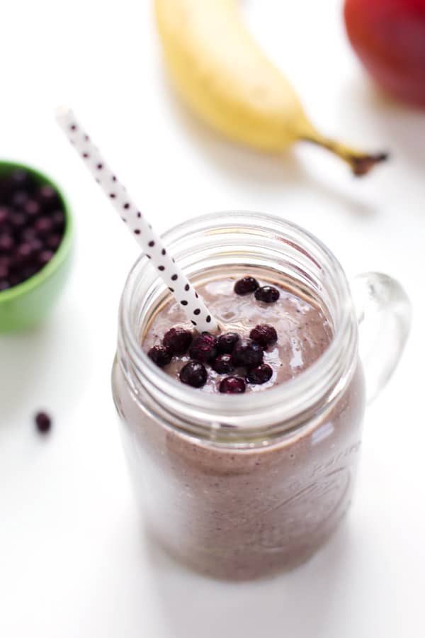 honey and wild blueberry smoothie in a glass mason jar topped with blueberries with a polka dotted straw in it on a white table with a green bowl of blueberries and a banana in the background