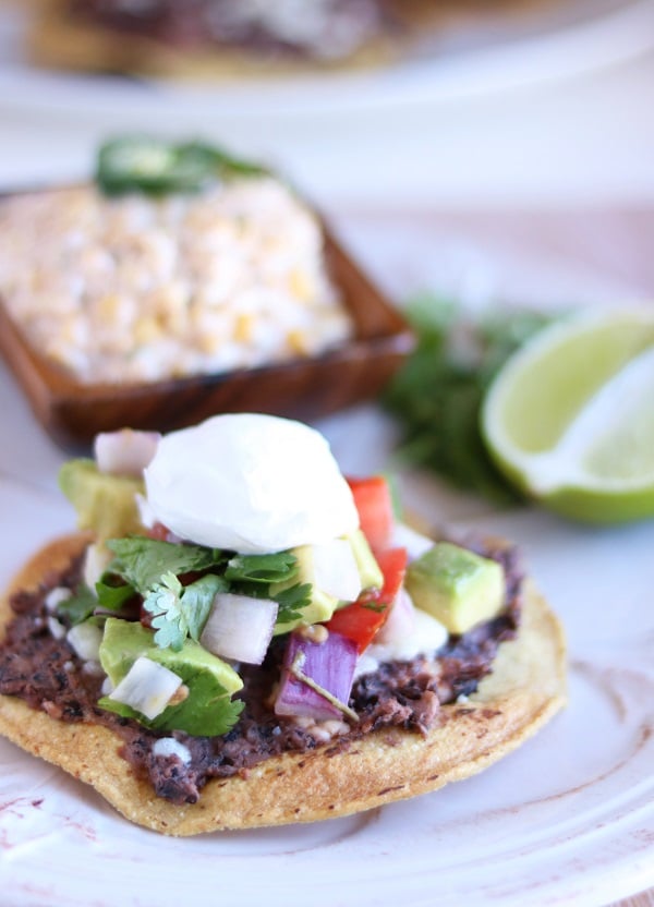 a black bean tostada topped with sour cream on a white plate with half a lime and a brown bowl of mexican corn salad in the background