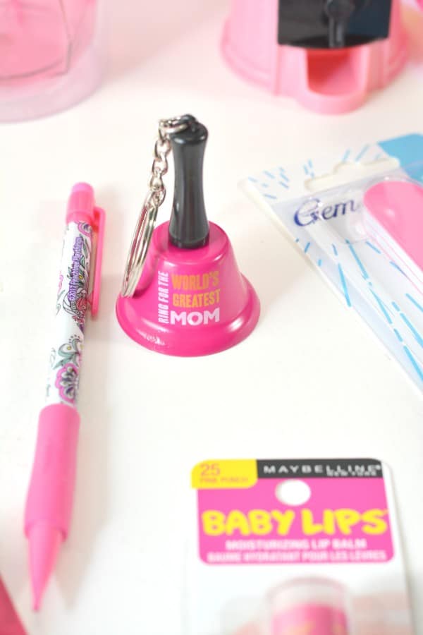 a pink pen, pink lip gloss, pink emery boards, pink bell with text on it reading World's Greatest Mom, all on a white table