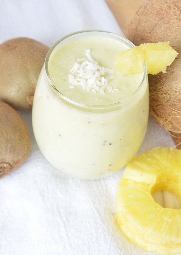 a Hawaiian tropical smoothie in a glass topped with shredded coconut and a pineapple slice on the edge, next to kiwis, a coconut and pineapple slices on a white background