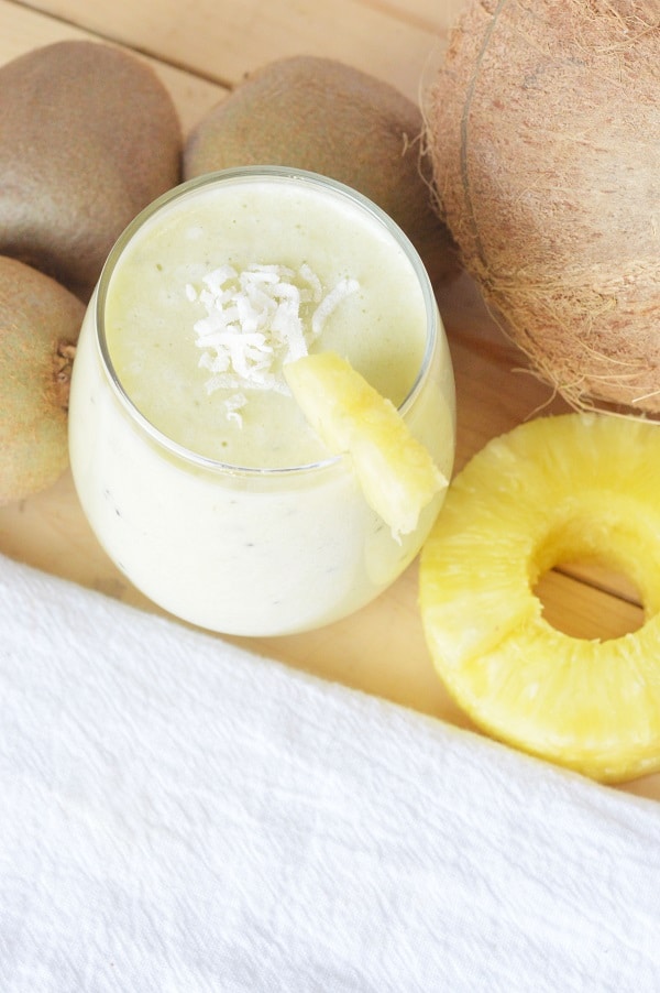 overhead view of a Hawaiian tropical smoothie in a glass topped with shredded coconut and a pineapple slice on the edge, next to kiwis, a coconut and pineapple slices on a white background