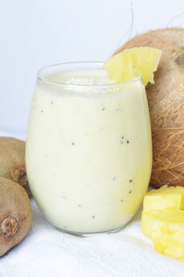 a Hawaiian tropical smoothie in a glass topped with shredded coconut and a pineapple slice on the edge, next to kiwis, a coconut and pineapple slices on a white background