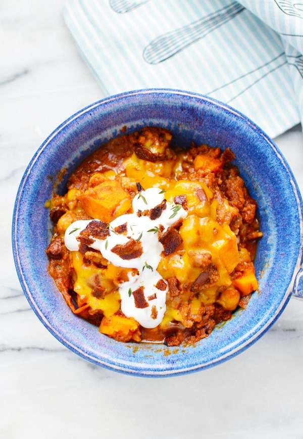 overhead view of sweet potato chili topped with sour cream and cheese in a blue bowl with a cloth with images of silverware on it in the background, all on a white kitchen counter