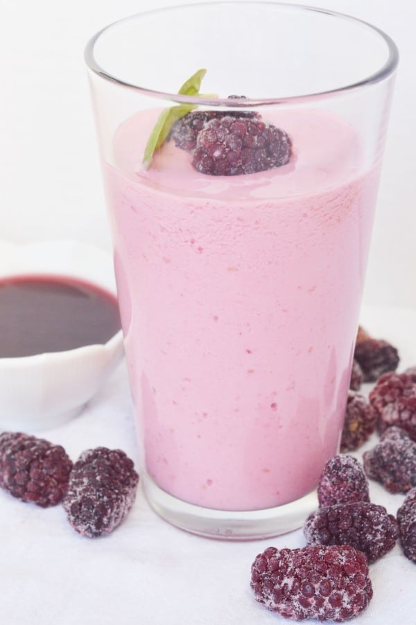 a raspberry cheesecake smoothie in a glass topped with two frozen raspberries and a green leaf surrounded by frozen raspberries and a white bowl of liquid raspberries on a white table 