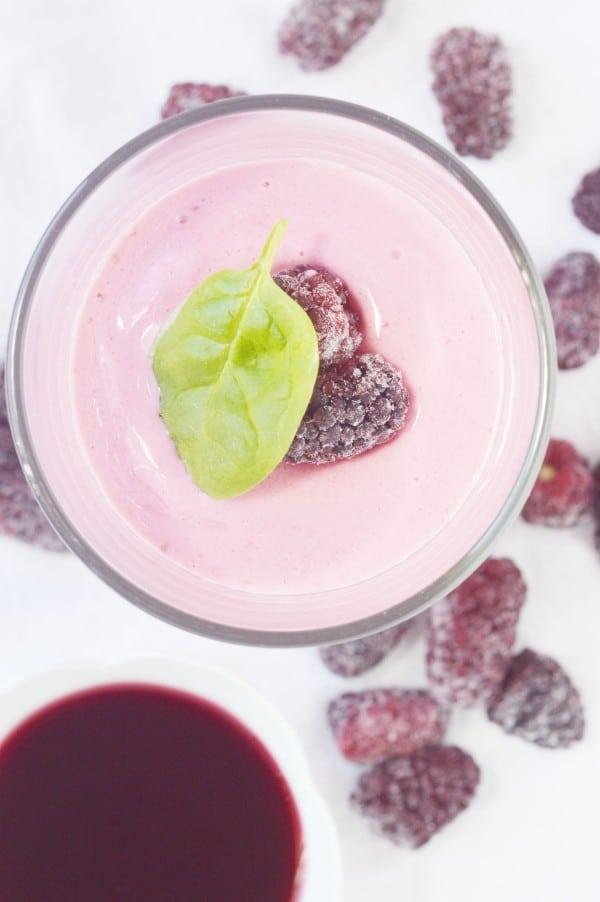 overhead view of a raspberry cheesecake smoothie in a glass topped with two frozen raspberries and a green leaf surrounded by frozen raspberries and a white bowl of liquid raspberries on a white table 