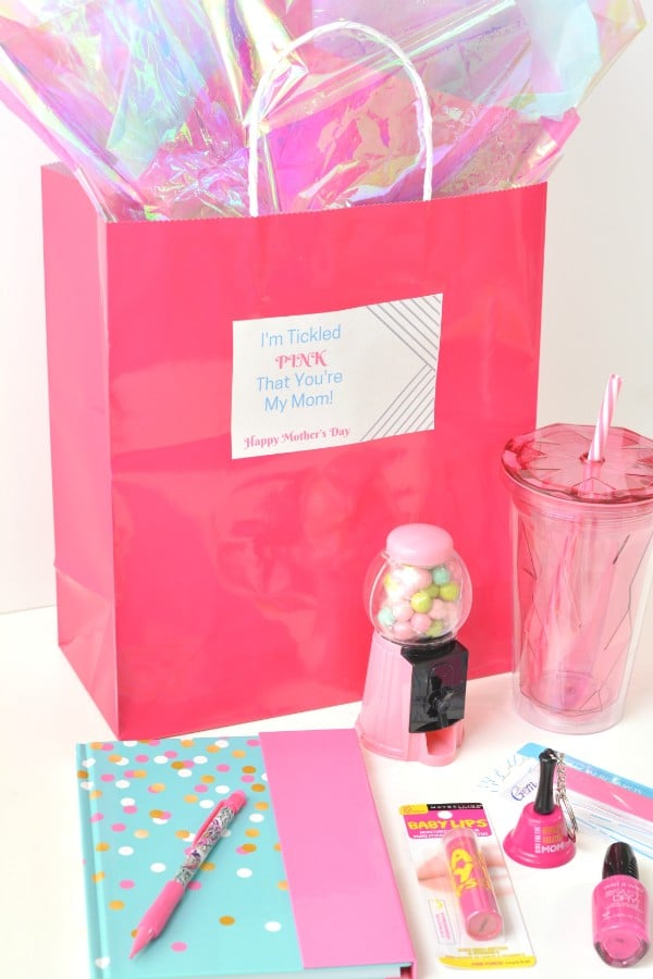 a pink bag with cellophane in it with a label on it reading, I'm tickled pink that you're my mom Happy Mothers Day, with a pink mini gumball machine, pink cup, pink notebook and pen, pink lip gloss, nail polish and an emery board in front of the bag, all on a white background