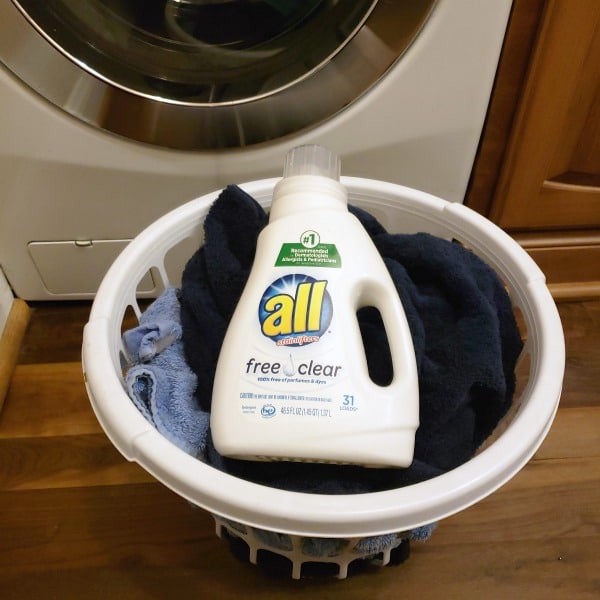 a basket of clothes with a bottle of All free and clear on top of it with a washer in the background