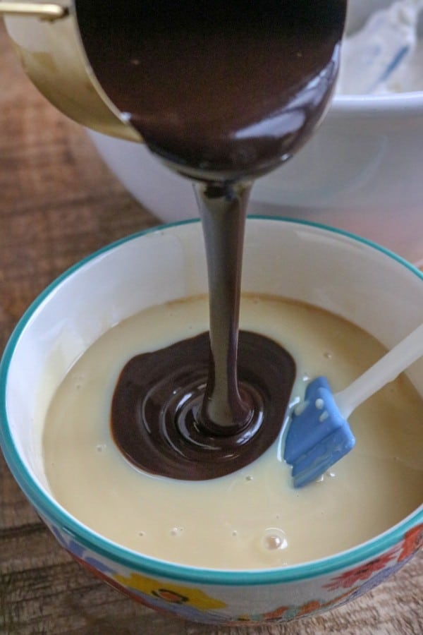 chocolate syrup being poured from a measuring cup into a bowl of sweetened condensed milk with a spatula in it next to a white bowl of whipped cream on a brown table