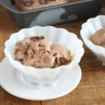 A white bowl filled with scoops of simple chocolate ice cream with chunks, placed on a matching white saucer. A tray with more ice cream is in the background.