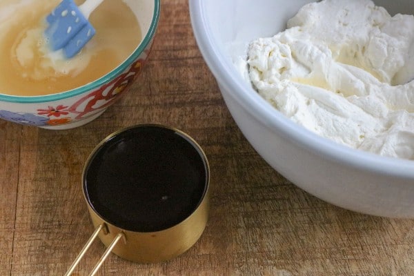 whipped cream and a spatula in a bowl of sweetened condensed milk next to a white bowl of whipped cream and a measuring cup of chocolate syrup on a brown table