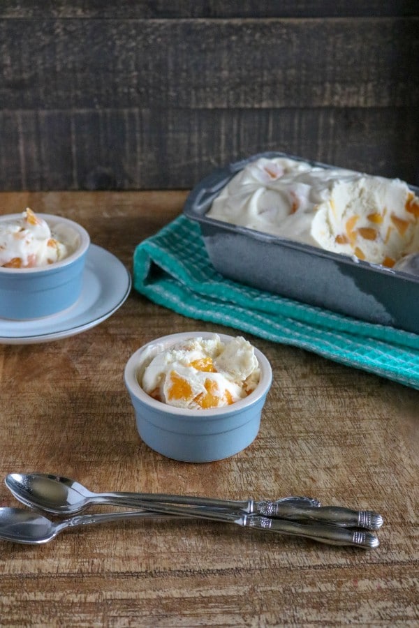 peach ice cream in blue bowls next to spoons with a metal pan on a green cloth in the background, all on a brown table