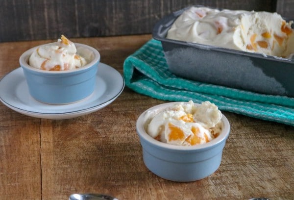 peach ice cream in two blue bowls, one on a saucer next to a metal pan of more peach ice cream on a green cloth on a brown table