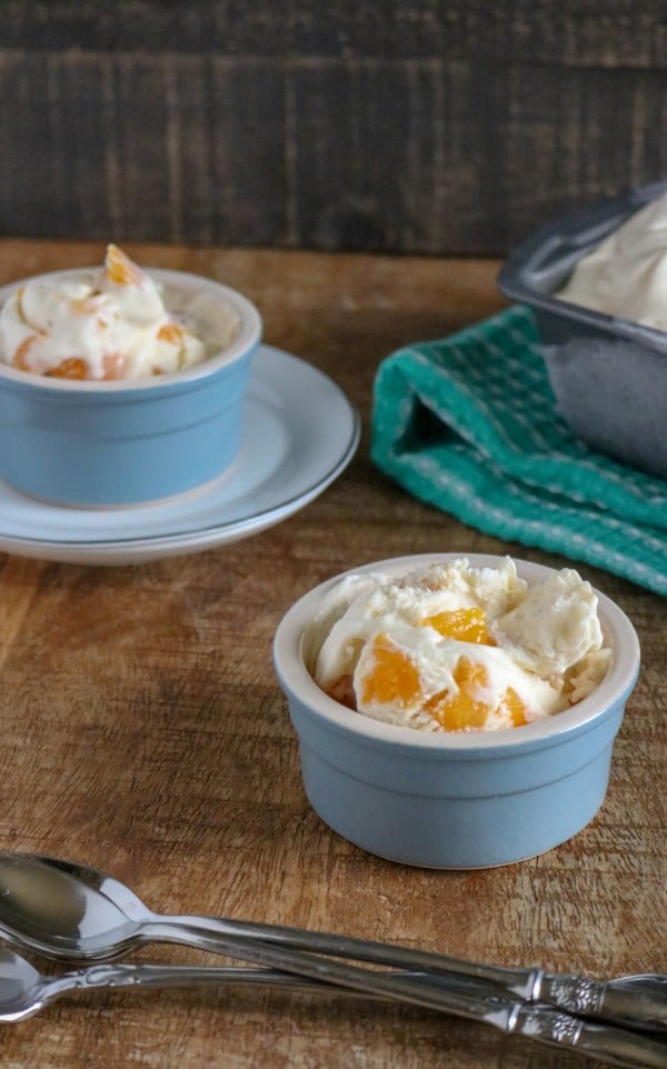 easy peach ice cream in two blue bowls next to spoons on a wood table with more ice cream in a pan in the background
