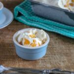 A small blue ramekin filled with easy peach ice cream and pieces of yellow fruit sits on a wooden surface next to a fork and spoon.