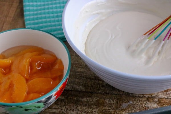 whipped cream and condensed milk in a white bowl next to a bowl of peach pie filling on a green cloth and brown table