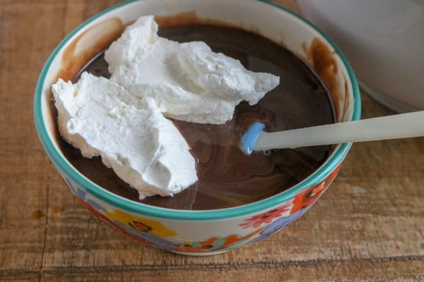 whipped cream on the top of the chocolate mixture in a bowl with a spatula in it on a brown table