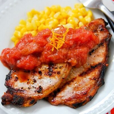 Grilled pork chops with rhubarb orange sauce, topped with zesty orange and served alongside corn on a white plate, offer a delicious twist on a classic dish.