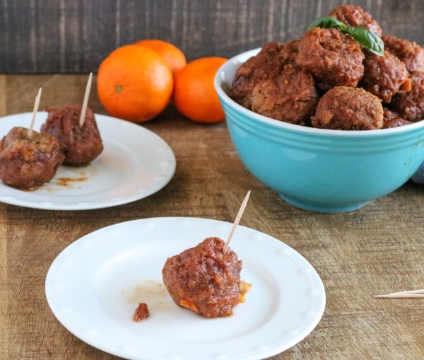 an orange glazed bbq meatball on a white plate next to another white plate with two meatballs on it next to a blue bowl full of meatballs on a brown table with oranges in the background
