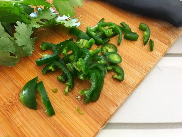 sliced jalapeno and cilantro leaves on a brown cutting board on a white wood table