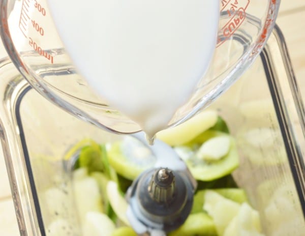 milk being poured into spinach leaves, kiwi and apple slices, and yogurt in a blender