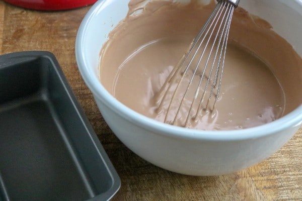 chocolate ice cream mixture in a white bowl with a wire whisk in it next to a metal pan on a brown table