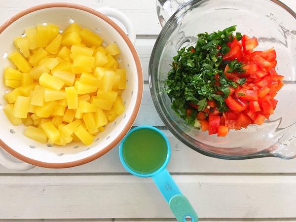 pineapple in a white colander, blue measuring cup with liquid in it, glass bowl with diced peppers and cilantro leaves on a white wood table