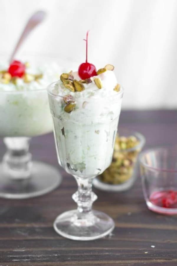 cool whip and pistachio salad topped with a cherry in glasses on a wood table