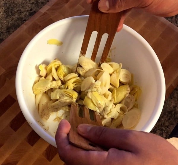 heart of palm and artichoke salad in a white bowl with a person tossing it using wooden salad forks