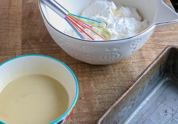 whipped cream in a white bowl with a multi-colored whisk, next to a bowl of sweetened condensed milk and a metal pan on a brown table